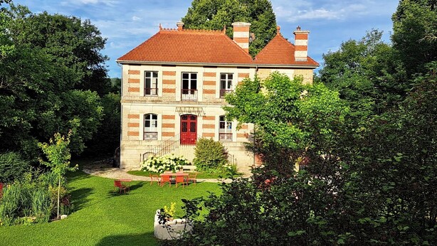 Una casa a due piani con una porta rossa, immersa nel verde e circondata da un prato curato.