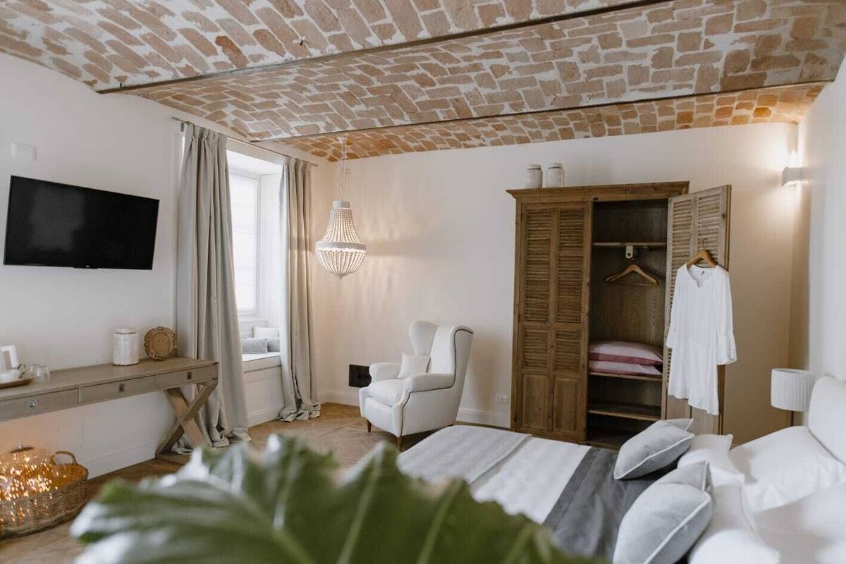 Quarto com teto de tijolos, uma cama, uma televisão, uma escrivaninha, uma cadeira, um guarda-roupa e um lustre.