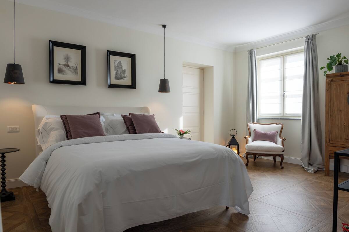 Quarto com uma cama grande, duas quadros na parede, uma cadeira, um armário e uma janela com cortinas.