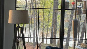 A room with a large window overlooking a forest and a body of water. There is a lamp, a chair, and a table with chess pieces visible. - Lake Hartwell water front with private dock, sleeps 15 (Hartwell)
