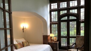 A bedroom with a large arched window, a bed with a white bedspread, a nightstand with a lamp, and a chair with a floral pattern. - Historic Traditional Estate - Heated Pool - Every Amenity (East Hampton)