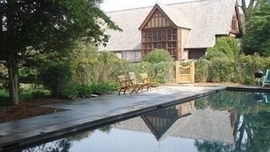 A swimming pool with a wooden deck and chairs, surrounded by lush greenery and a house with a gabled roof. - Historic Traditional Estate - Heated Pool - Every Amenity (East Hampton)