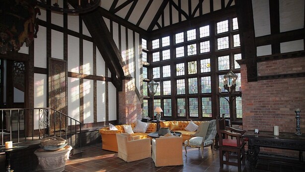 A spacious room with a high ceiling, exposed wooden beams, and large windows allowing natural light. There is a seating area with sofas and a table, and a staircase leading to an upper level. - Historic Traditional Estate - Heated Pool - Every Amenity (East Hampton)