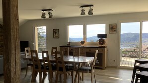 A dining area with a wooden table and chairs, a kitchen area with a window, and a view of the outdoors. - New house for 2 to 8 people at the foot of the slopes with panoramic view of the lake (Gérardmer)