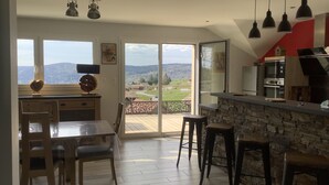 A modern kitchen with a large window, a dining area with wooden chairs, and a stone accent wall. - New house for 2 to 8 people at the foot of the slopes with panoramic view of the lake (Gérardmer)
