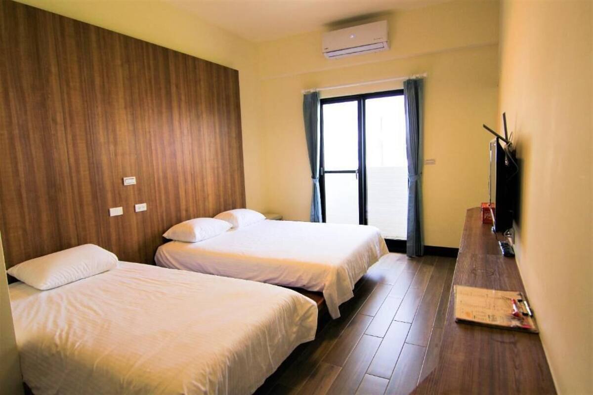 A hotel room with two beds, a wooden panel wall, a large window with curtains, and a flat-screen TV on a wooden table.
