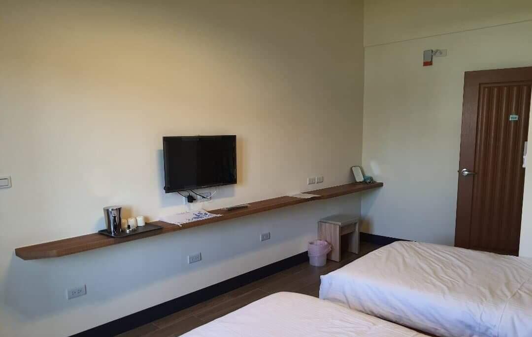 A hotel room with a bed, a TV mounted on a wooden shelf, a small table, and a door.