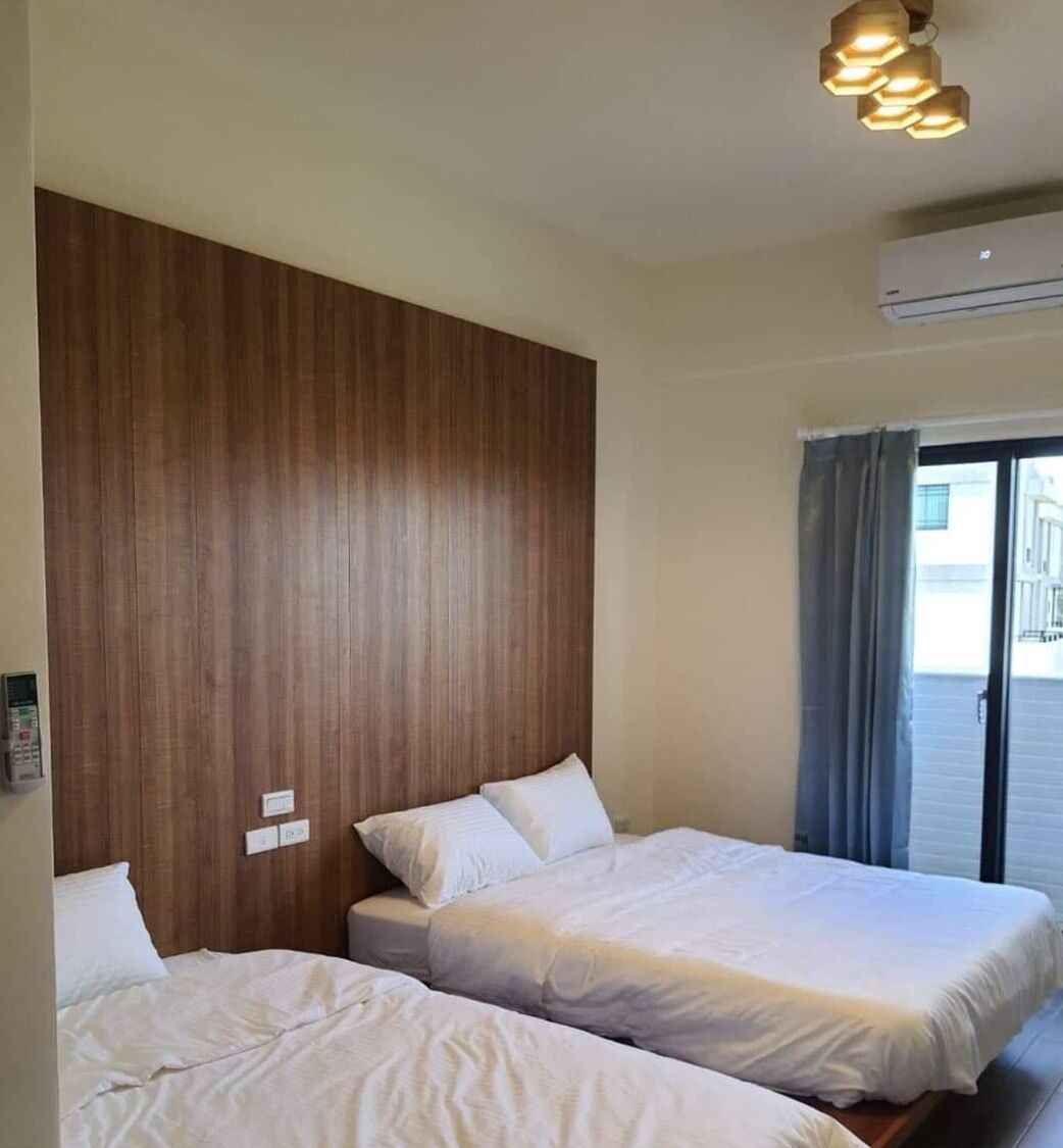 A hotel room with two beds, wooden paneling on the wall, an air conditioning unit, and a view of the outside through a window with curtains.
