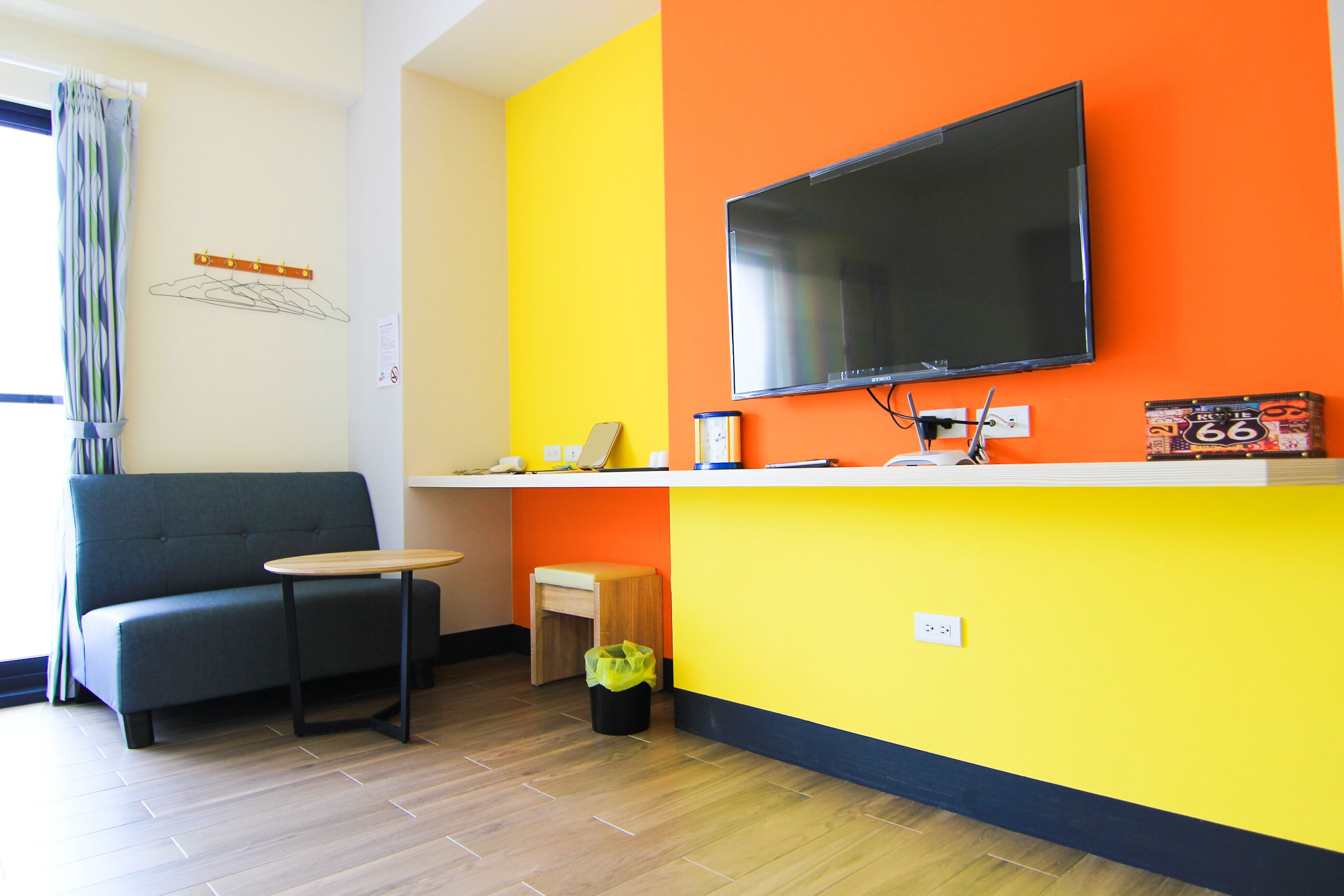 A modern living room with a sofa, a small table, a TV mounted on a yellow wall, and a small wooden stool.
