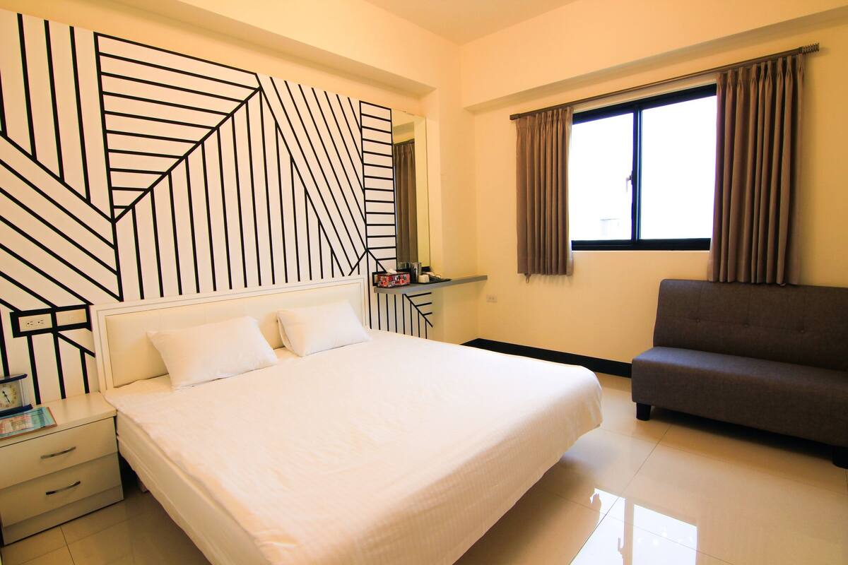 A hotel room with a large bed, a grey sofa, a desk with a mirror, and a window with curtains.