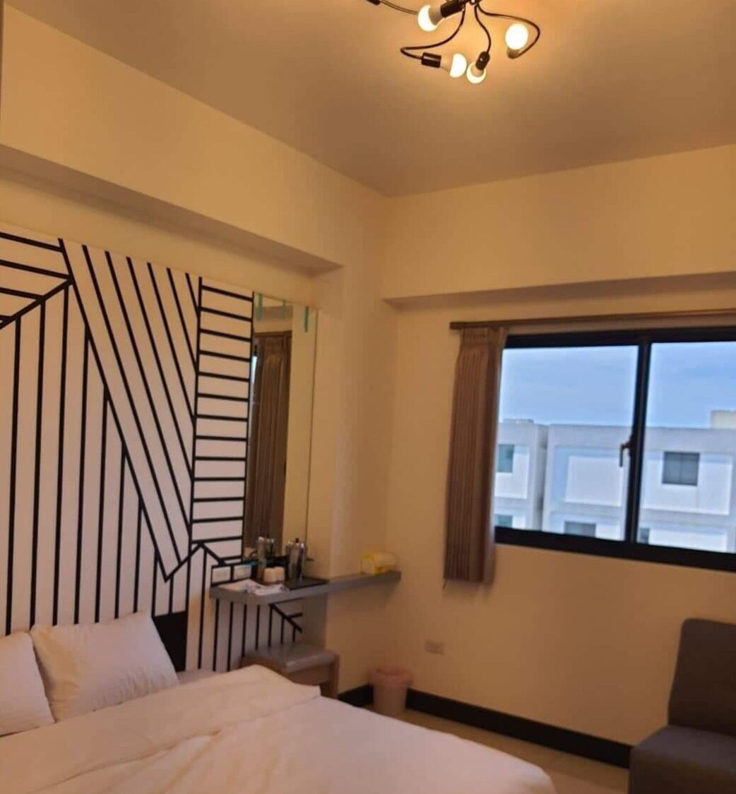 Une chambre d’hôtel comprenant un lit, un bureau avec une lampe, une fenêtre avec des rideaux et un panneau décoratif mural.