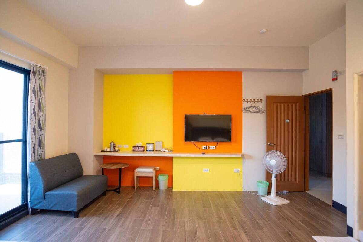 A room with a sofa, a small table, a TV mounted on an orange wall, a fan, and a wooden door.