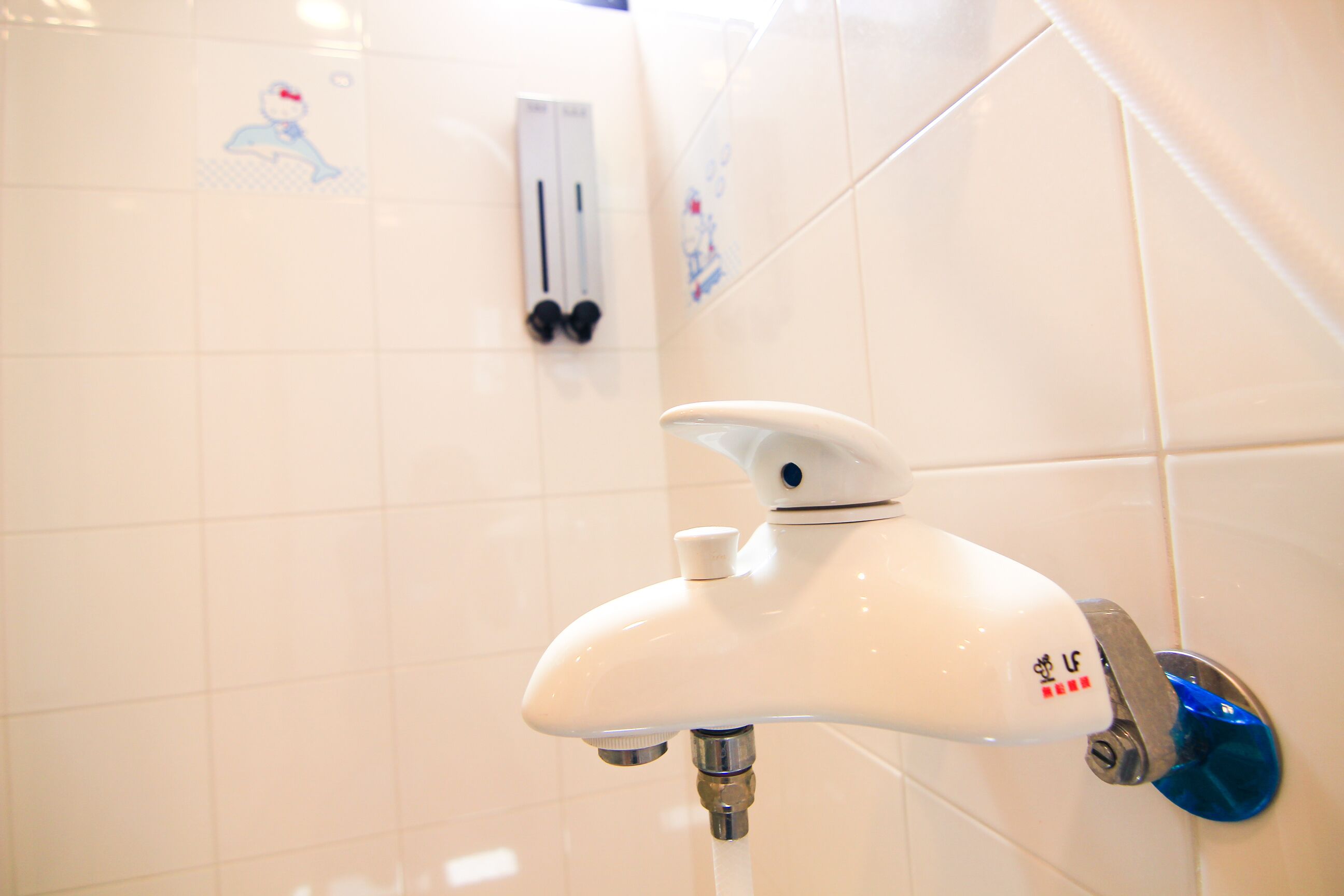 A bathroom with white tiled walls, a white faucet with a blue handle, and a soap dispenser on the wall.