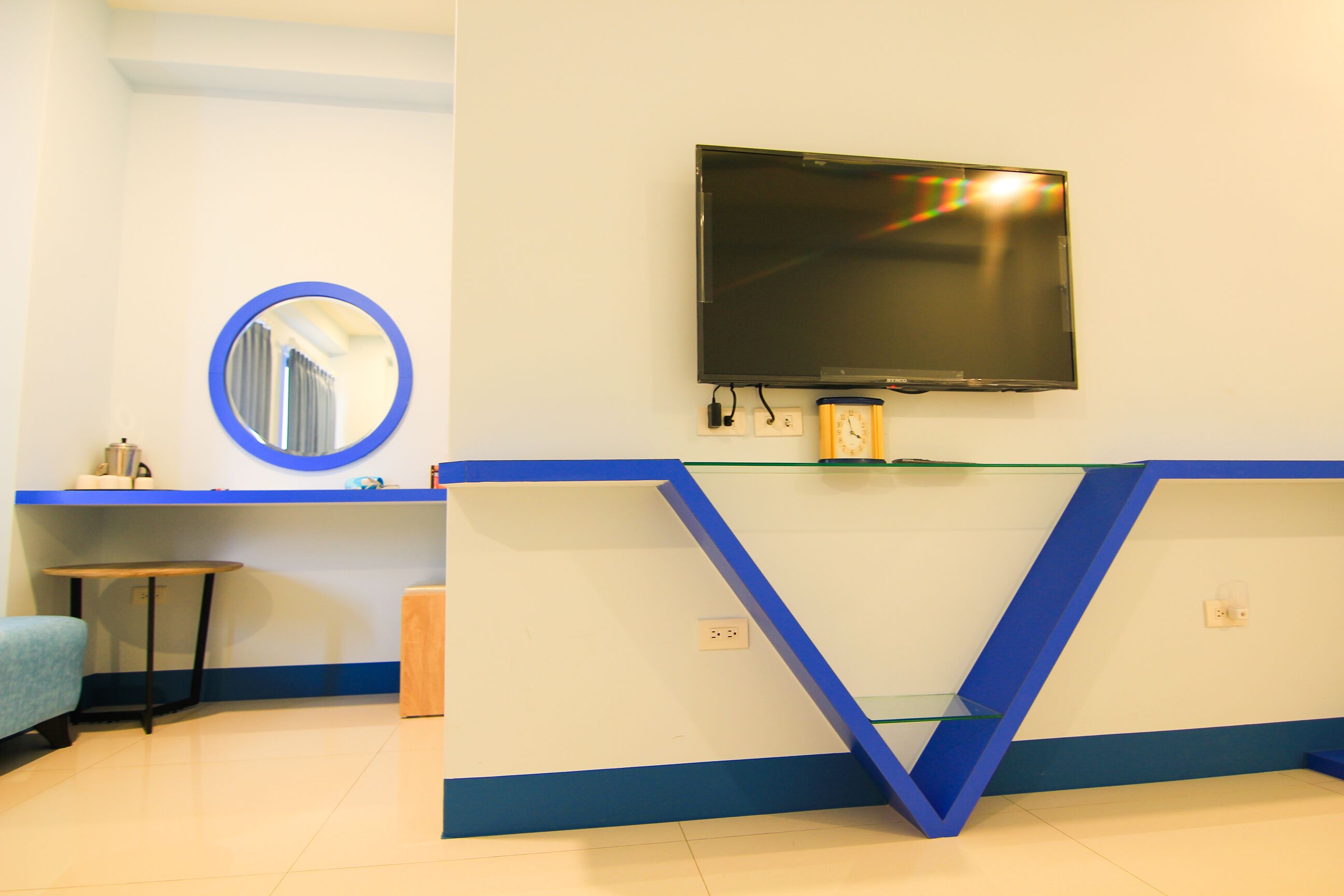 A room with a flat-screen TV mounted on the wall, a round mirror, and a blue-framed glass shelf.