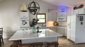 Una cocina moderna con armarios blancos, una isla grande con taburetes, una lámpara colgante y un letrero de neón que dice "Paddle".