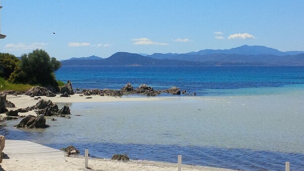 A beach with clear blue water, rocky shoreline, and mountains in the background.