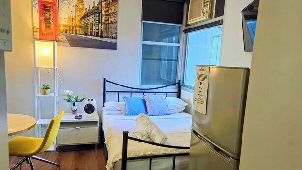 A compact hotel room with a bed, a desk with a lamp, a chair, a small table, a refrigerator, and a wall-mounted air conditioner. - studio on  Swanston st apartment with a little English flare (Carlton)
