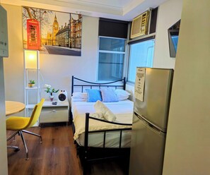 Une chambre d’hôtel compacte comprenant un lit, un bureau avec une lampe, une chaise, une petite table, un réfrigérateur et un climatiseur fixé au mur.