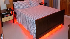 Ett sovrum med en säng, ett nattduksbord, en lampa, en takfläkt och LED-belysning längs sängens fotgavel.
