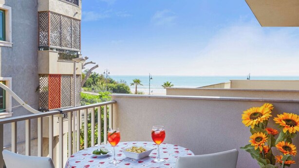 A balcony with a table set for two, two drinks, a bowl of snacks, and a potted plant with sunflowers. - Comfortable apartment  (Pozzallo)