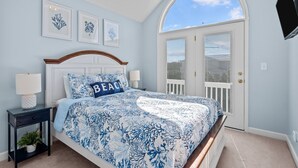 A bedroom with a bed, a nightstand, a potted plant, and a door with a view. - Andreas Place Spacious Family Getaway, Easy Beach Access, Pool (Kure Beach)
