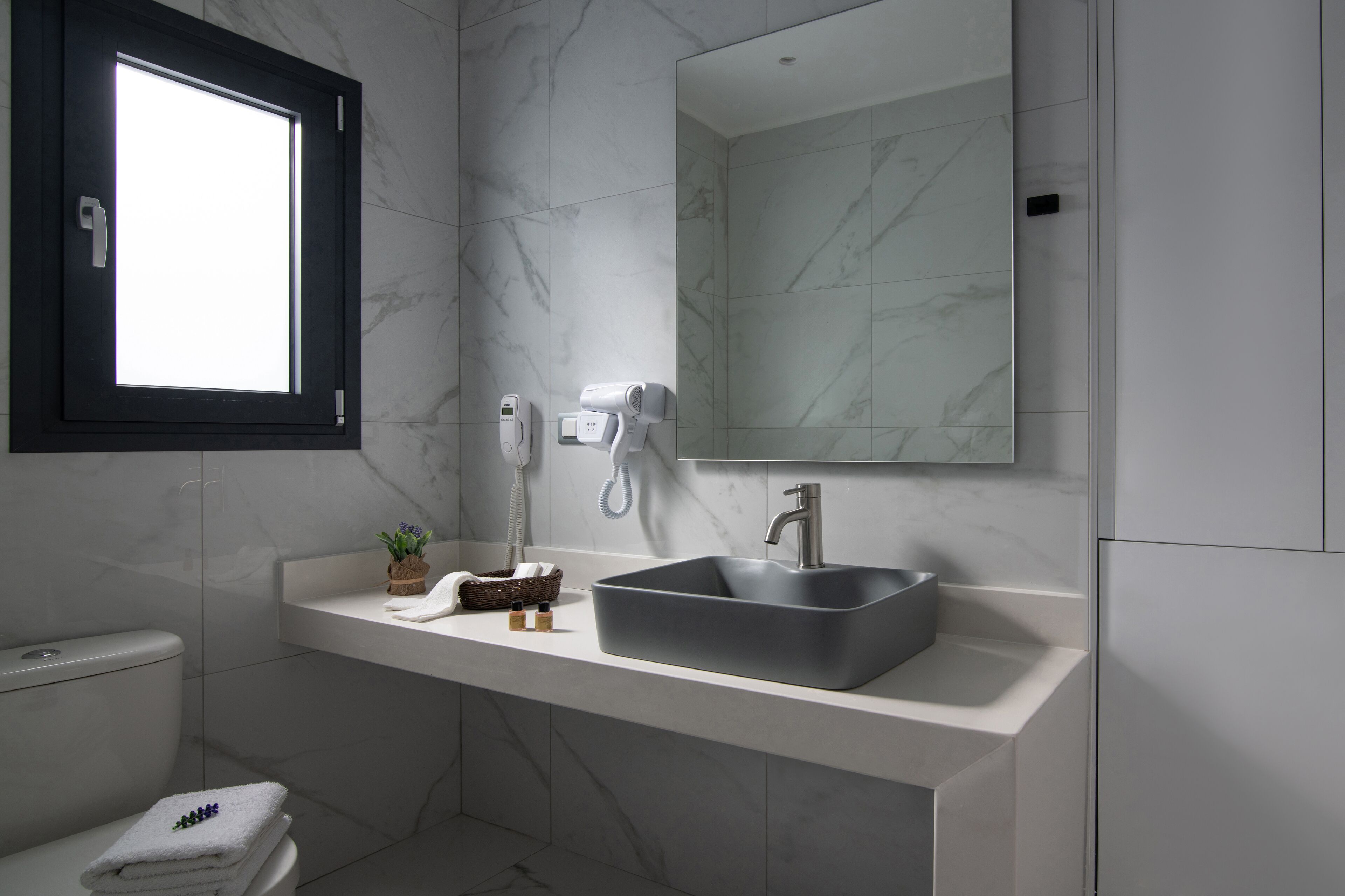 Ein modernes Badezimmer mit Marmorwand, großem Spiegel, Waschbecken, Toilette und Fenster.
