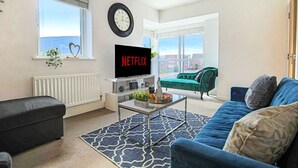 A living room with a television, a sofa, a coffee table, and a window with a view of buildings. - 🌟Large house with Free Parking, Smart TV & Garden🌟 (Bletchley)