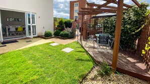 A backyard with a wooden pergola, a patio table, and chairs. There is a grassy area with stepping stones and a brick wall. - 🌟Large house with Free Parking, Smart TV & Garden🌟 (Bletchley)