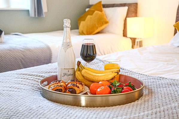 A breakfast tray with croissants, bananas, tomatoes, strawberries, a bottle of Freixenet ice wine, and a glass of red wine on a bed.
