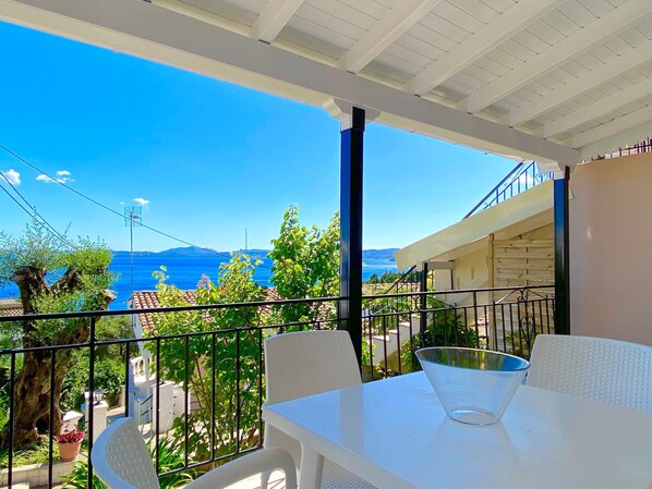 A balcony with a clear view of the sea and mountains, white furniture, and a metal railing.
