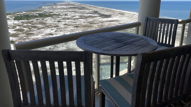 Ein Balkon mit rundem Tisch und zwei Holzstühlen, mit Blick auf einen Strand und klare Sicht aufs Meer.