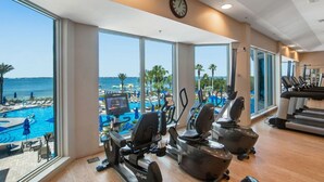 Ein Fitnessraum mit Cardiogeräten, einer Uhr und Blick auf Pool und Strand.