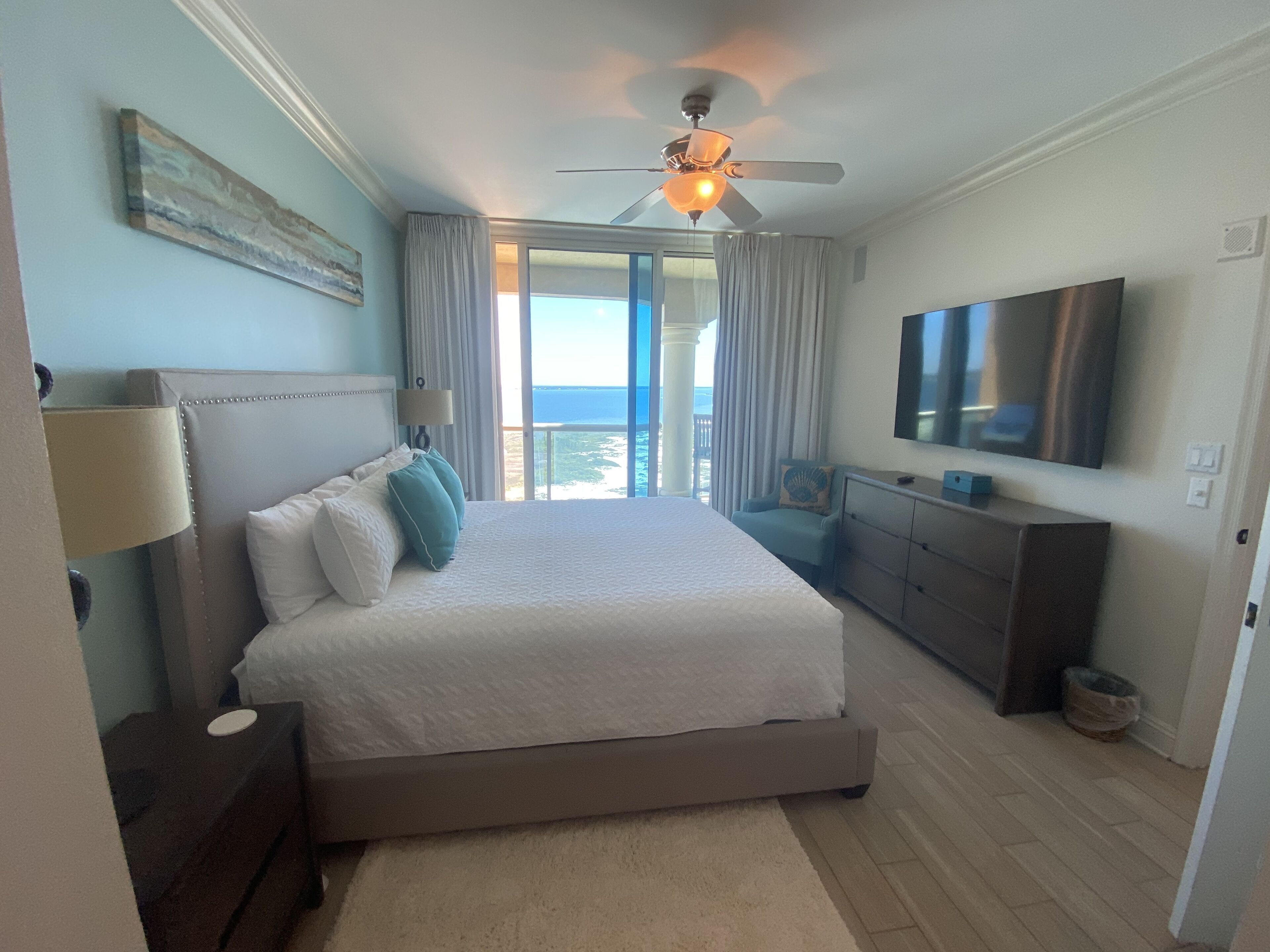 Un dormitorio con cama, ventilador de techo, televisión, cómoda y vista al océano.