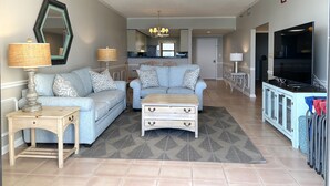 Ein Wohnzimmer mit Sofa, Couchtisch und TV-Standfuß, im Hintergrund eine Küche und ein Deckenventilator.