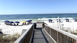 Ein Holzsteg führt zu einem Strand mit Sonnenschirmen und Liegestühlen.