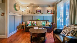 A cozy living room with a wooden coffee table, a sofa with floral cushions, and a window with sheer curtains.