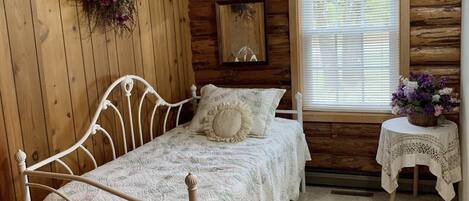 A cozy cabin room with a metal bed, wooden walls, a window with blinds, a small table with a vase, and a wall-mounted mirror.