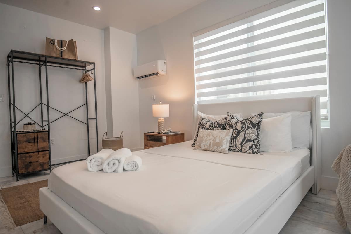 Una cama bien hecha con toallas enrolladas, un armario de madera, una mesita de noche con lámpara y una ventana con persianas.
