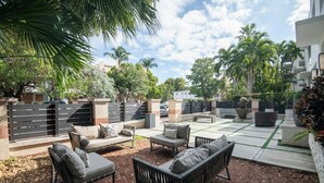 A patio area with outdoor furniture, including sofas and a table, surrounded by a low brick wall and palm trees. - Renzzi On The Beach (Miami Beach)