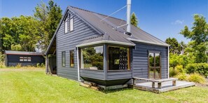 Ett litet, enplanshus med en mörkgrå fasad, ett sluttande tak och en skorsten. Det har ett stort fönster på framsidan och en liten veranda med en bänk.