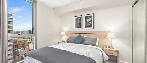 A hotel room with a large bed, two bedside tables with lamps, a view of the city through a large window, and two framed pictures on the wall.