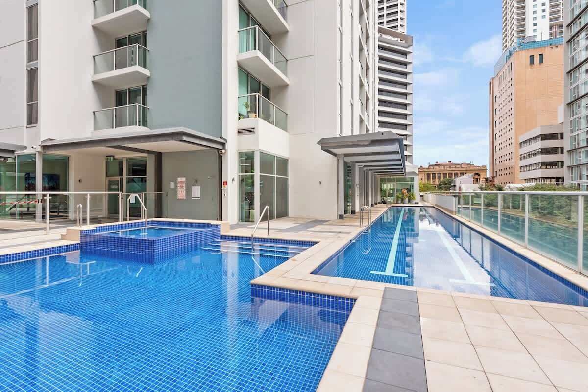 Ein Schwimmbeckenbereich mit mehreren Pools, umgeben von modernen Gebäuden mit Balkonen und großen Fenstern.