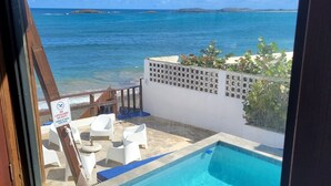On peut voir une piscine avec une terrasse en bois et des chaises longues, un panneau indiquant qu’il est interdit de plonger, ainsi qu’un océan bleu clair avec des îles à l’horizon.