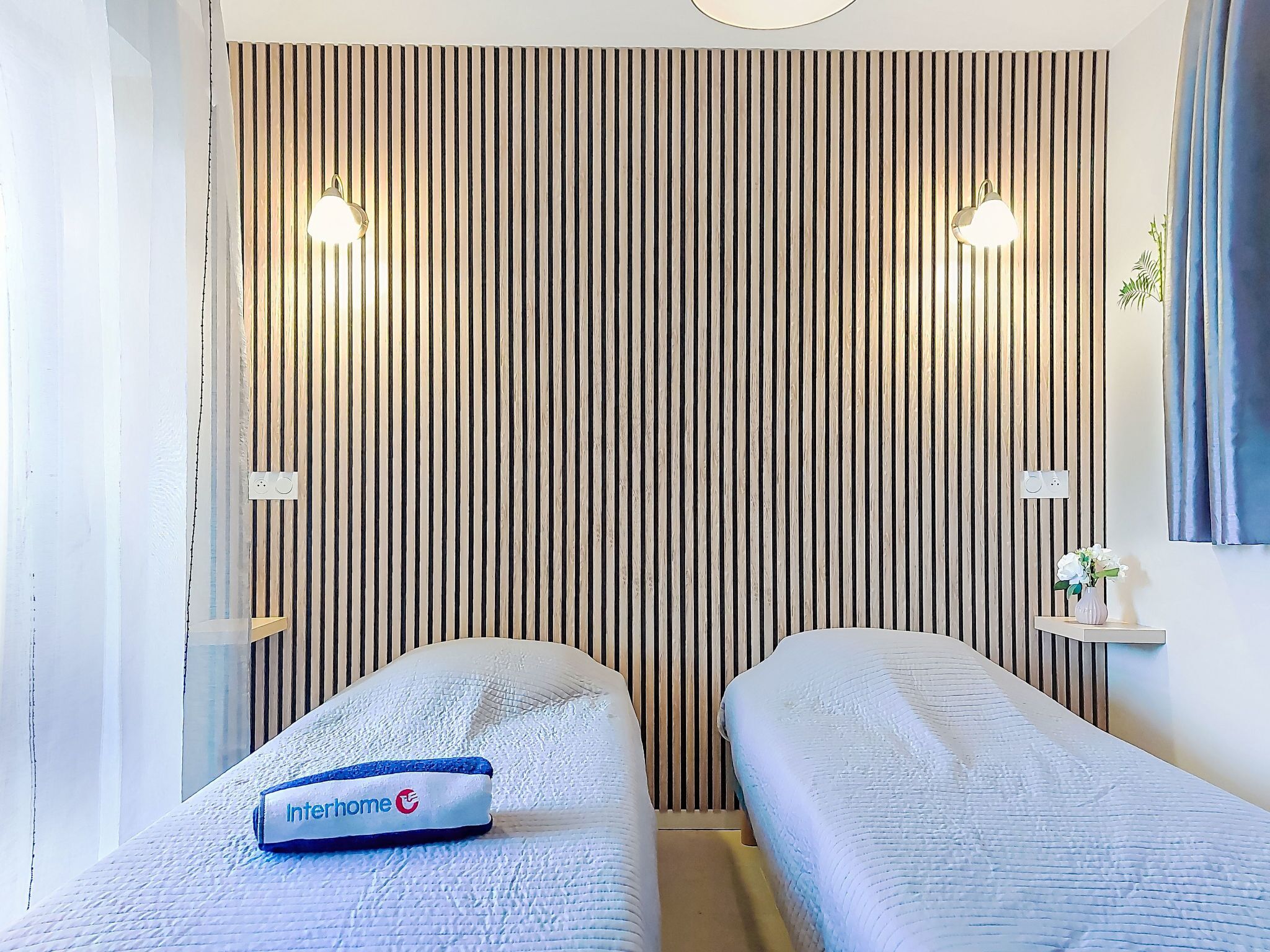 A hotel room with a striped wall, two beds, bedside lamps, a small table with a vase, and a sign on the bed.