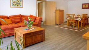 Una sala de estar con sofá, mesa de centro, mesa de comedor con sillas, un aparador y una puerta que da a otra habitación.