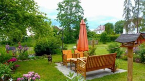 Un patio jardín con mobiliario de madera y una sombrilla, rodeado de abundante vegetación y flores.
