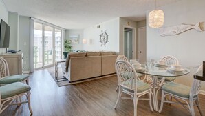 A spacious living area with a sofa, dining table, and chairs. There is a television on the left, a large window, and a door leading to another room. - 🌊Labor Day Available! Aug 30-Sept 3🔥Oceanfront🏝️2 Pools🐬Restaurants Onsite☀️ (North Topsail Beach)