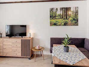 A living room with a TV on a wooden cabinet, a small round table with a plant, a sofa, and a forest-themed wall art.