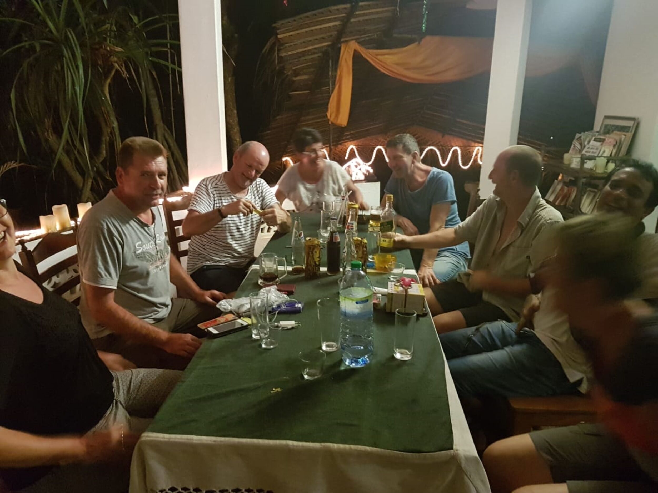 Un grupo de personas sentadas alrededor de una mesa con varias botellas y vasos, en una habitación con techo de madera y plantas al fondo.