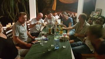 Un grupo de personas sentadas alrededor de una mesa con varias botellas y vasos, en una habitación con techo de madera y plantas al fondo.